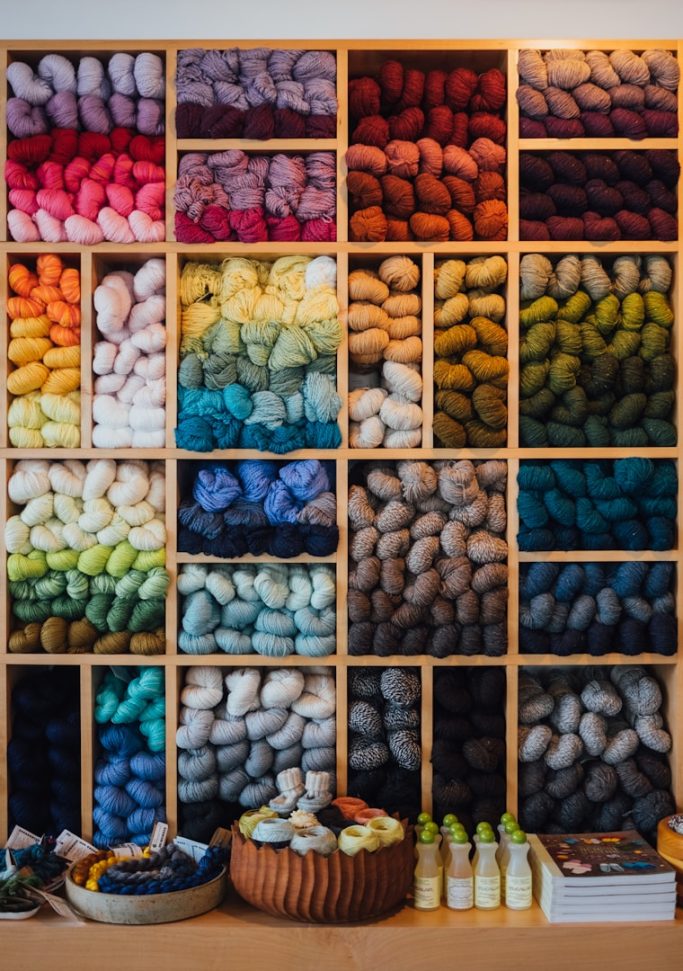 Wolle Bunte Wolle in Regalen, geordnet nach Farben von Rot bis Blau und einer Auswahl an Zubehör.